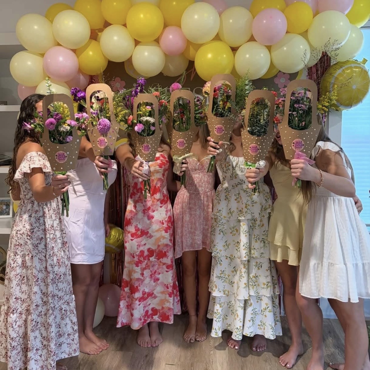 Group of children holding decorated signs in front of yellow, pink, and white balloons, celebrating a special occasion.