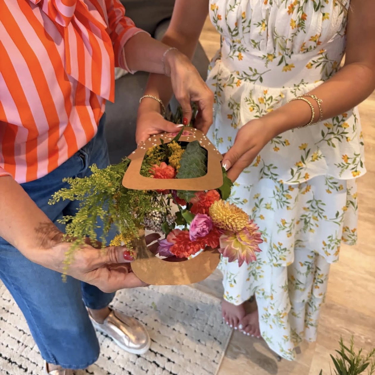 Two women exchange a gift box with flowers and greenery inside, one wearing a striped orange and white top, the other in a floral dress.