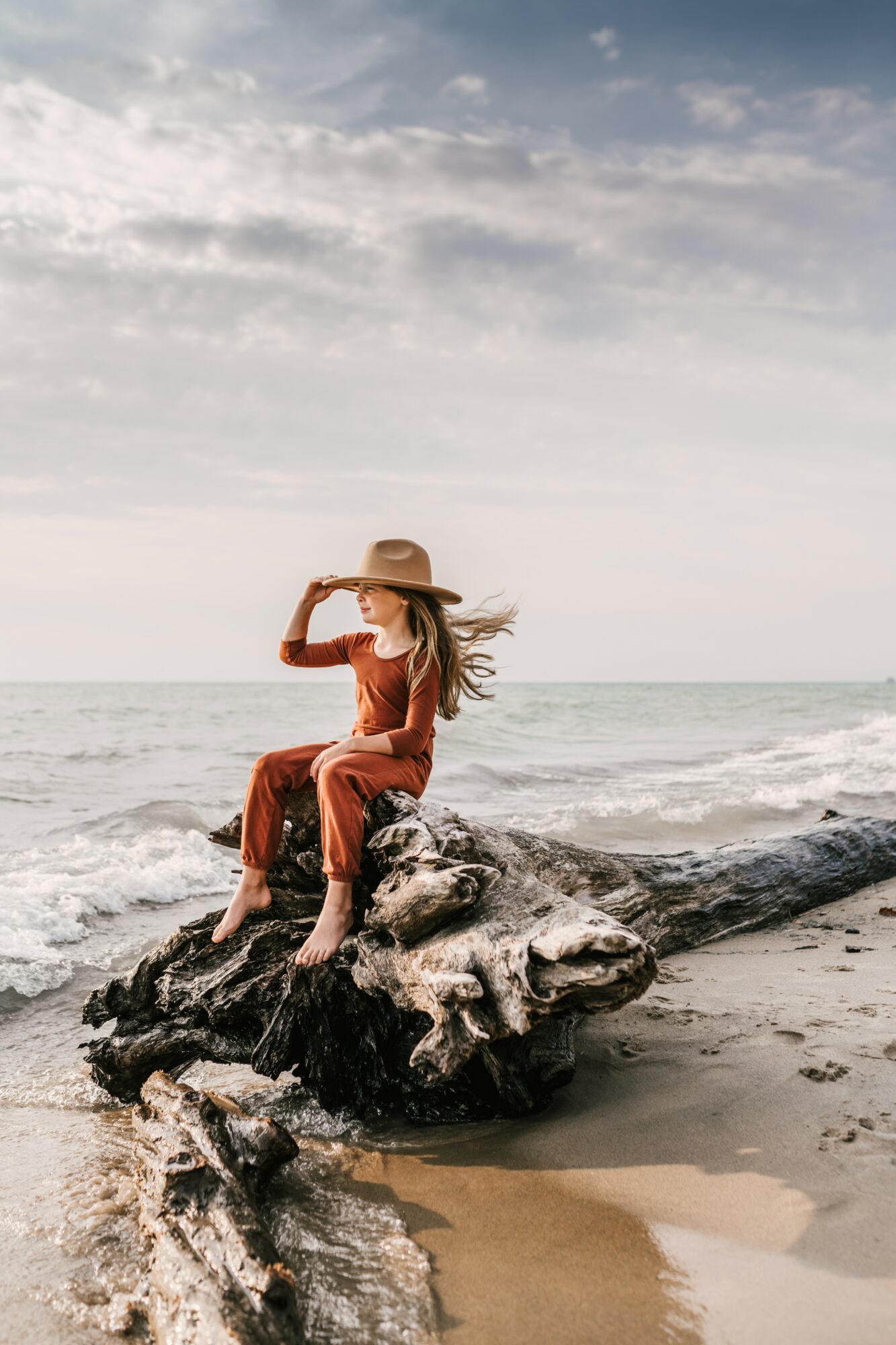 Woman in a long dress and wide-brimmed hat sitting on a driftwood log at the beach, looking at the ocean, cloudy sky.