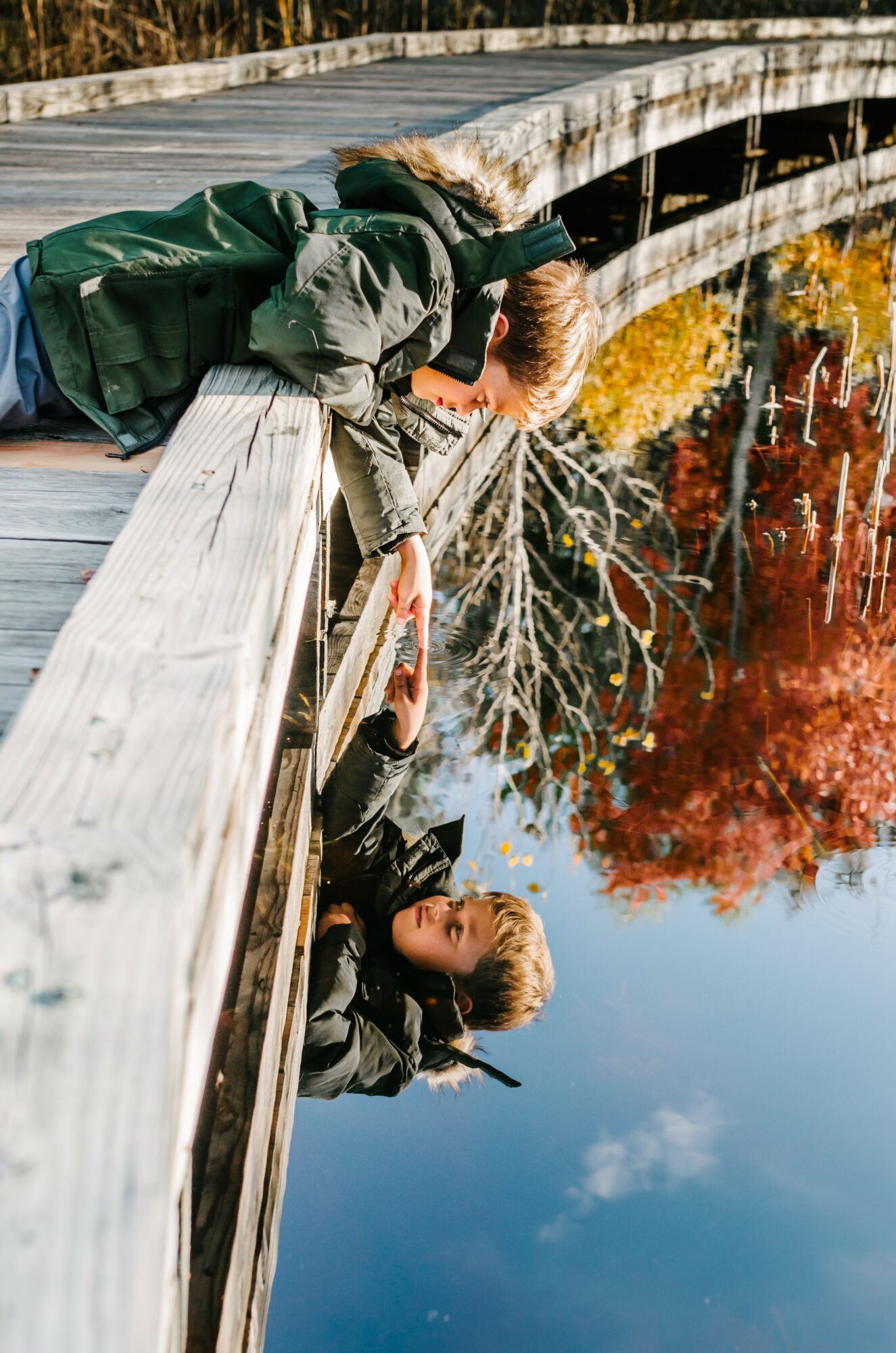 Two children leaning over a wooden bridge, looking at water with colorful trees in the background.