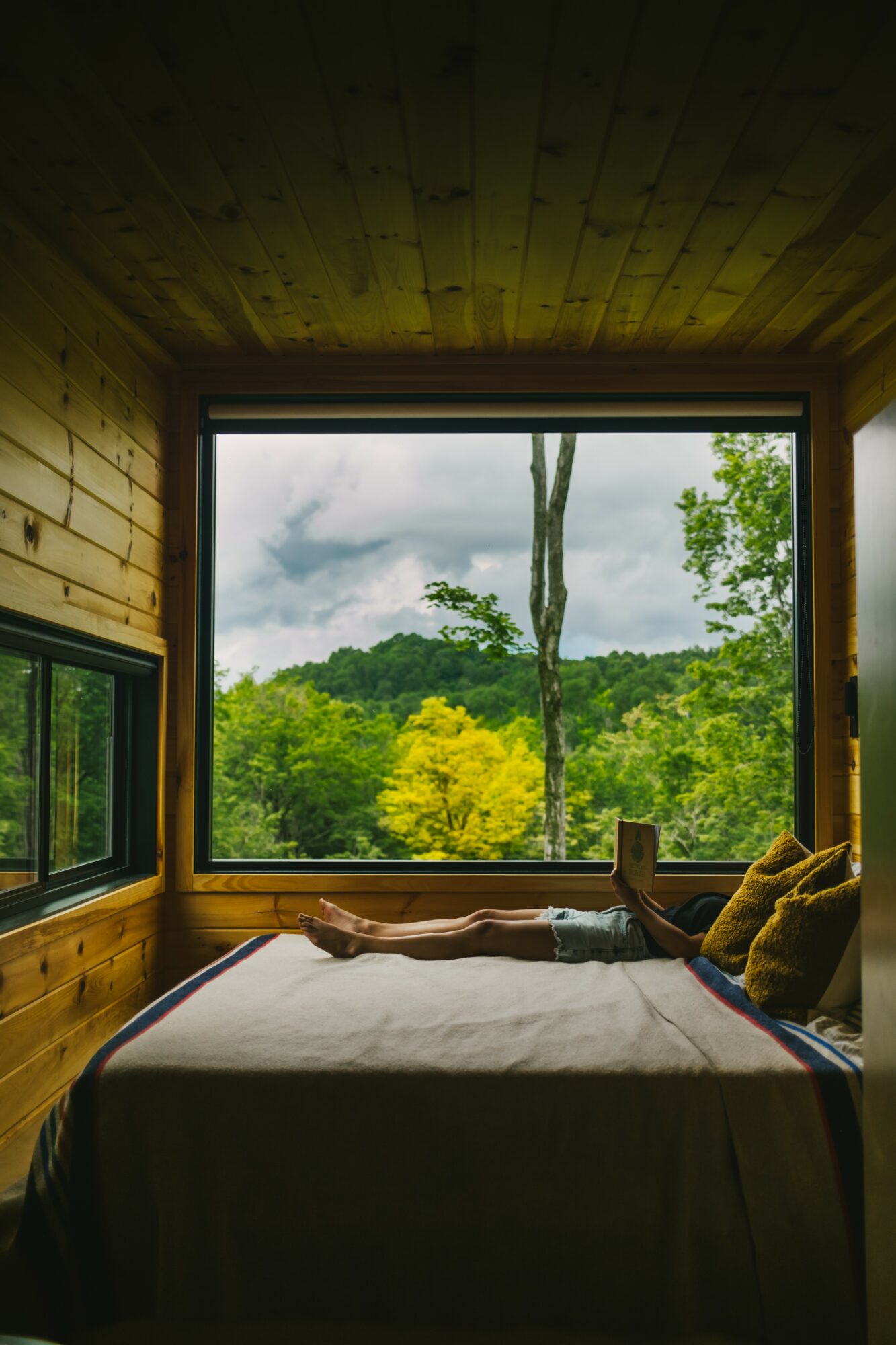 Person lying on bed inside wooden cabin, looking out large window at green trees and cloudy sky.