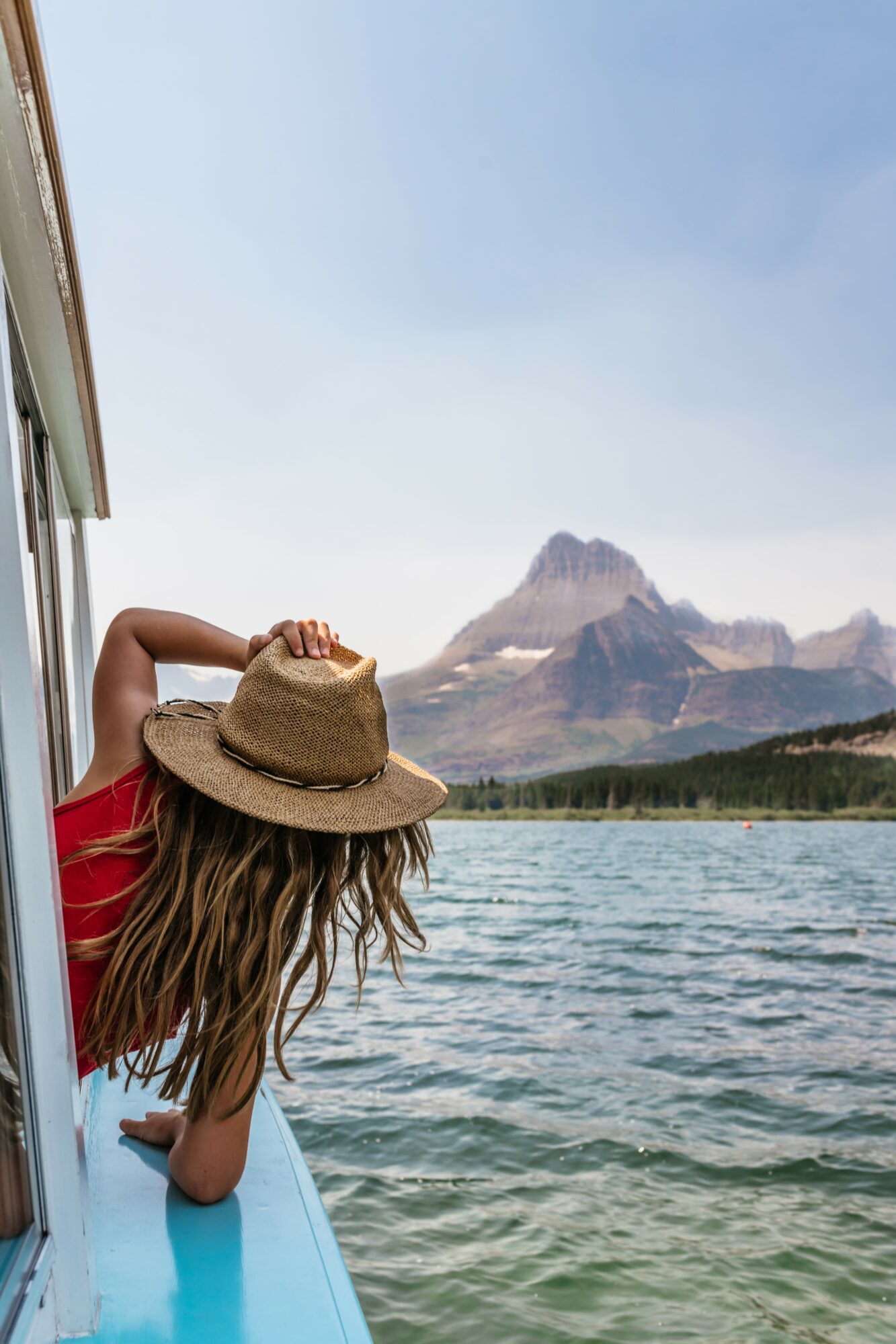 Person with long hair and a hat leaning out of a boat, mountain and water in background.