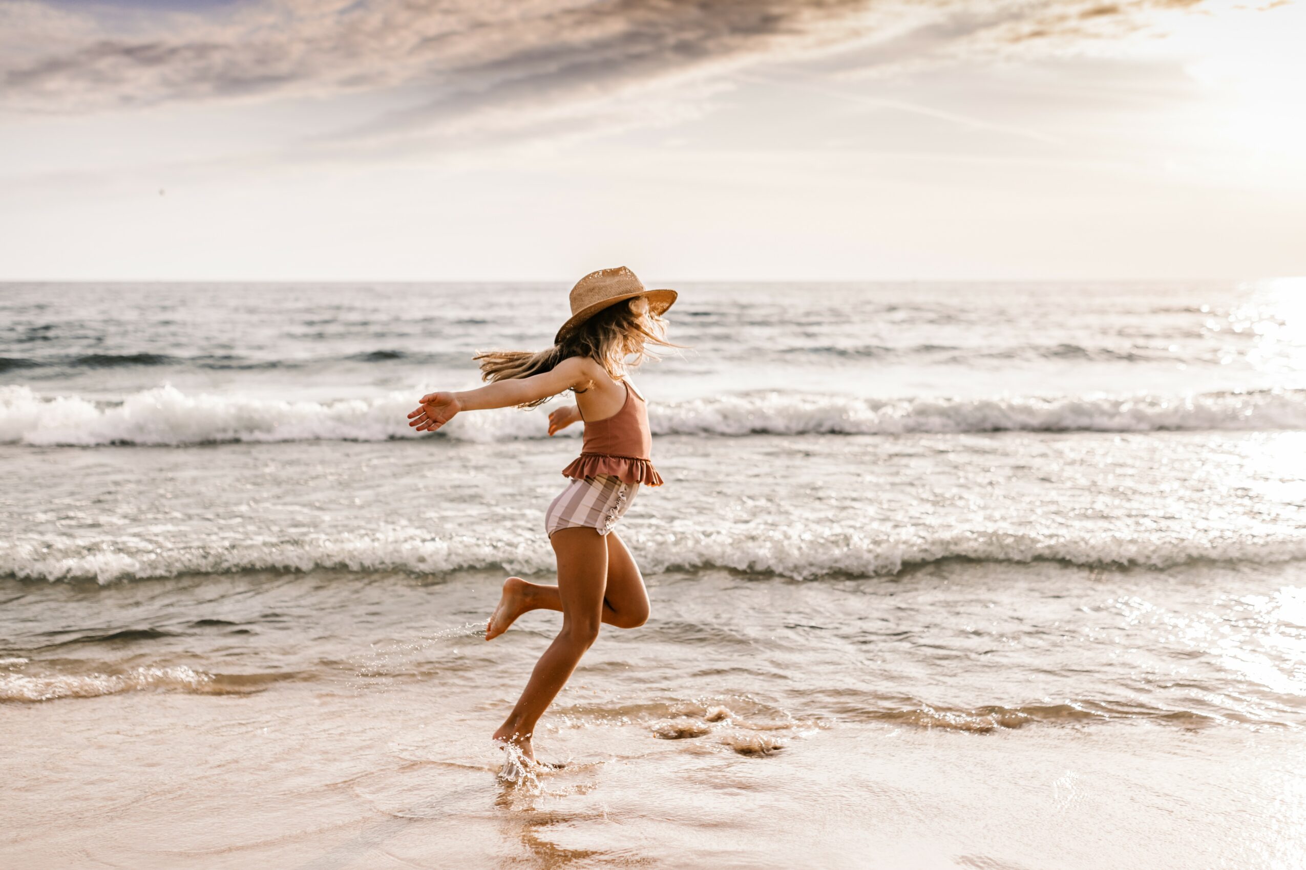 Girl in a hat and swimsuit running in shallow ocean water at the beach, waves and sky in background.