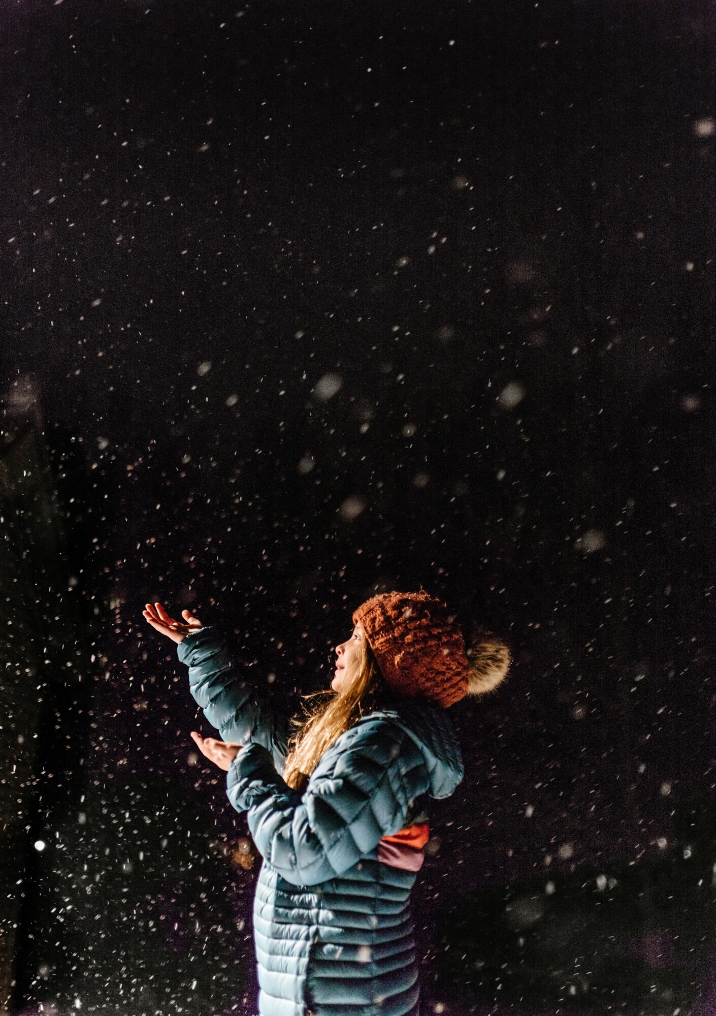 Child in winter coat and hat reaching up in falling snow at night, dark background