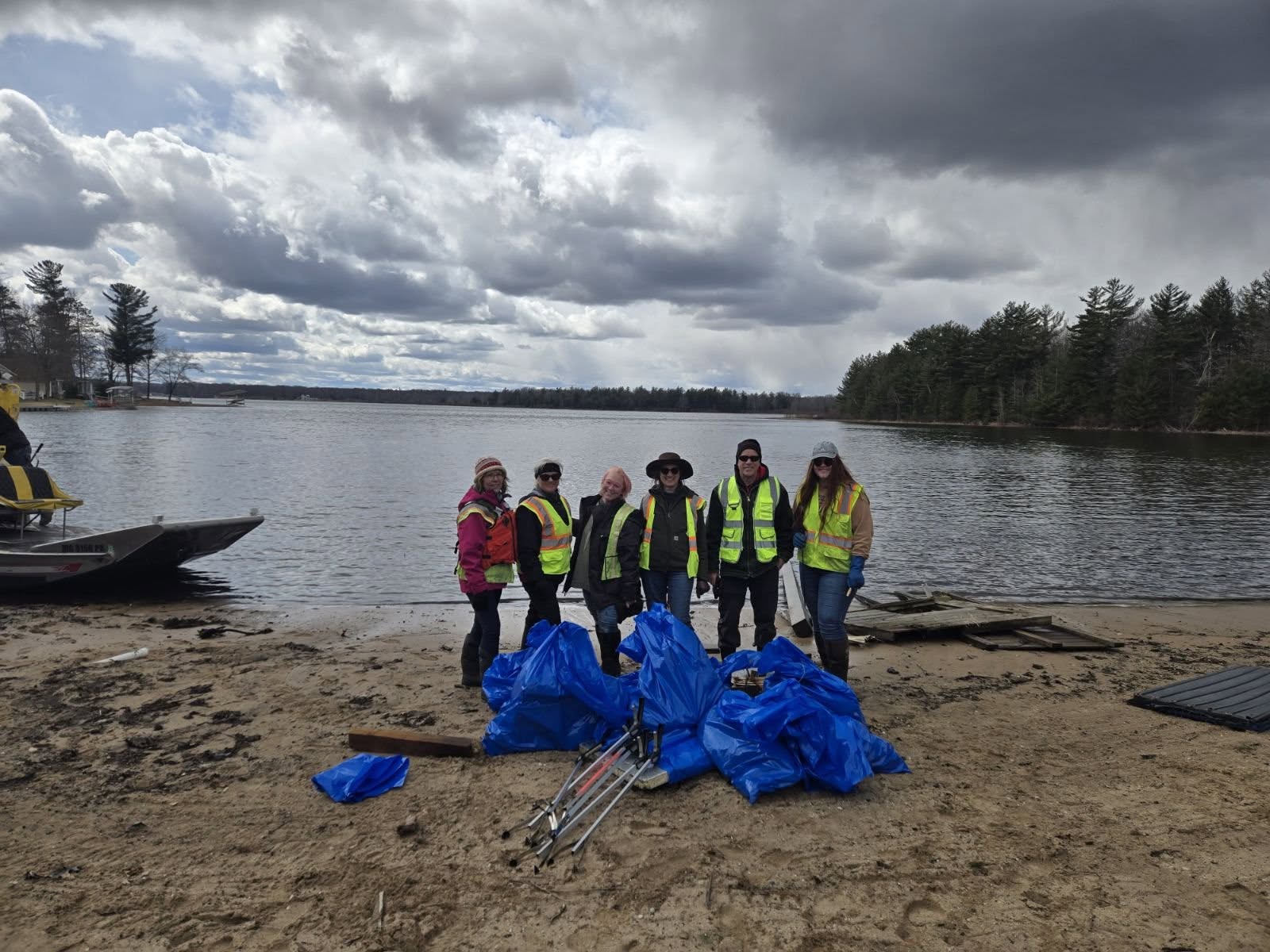 Group of people wearing yellow safety vests on a beach near a lake, with blue bags and equipment, cloudy sky.