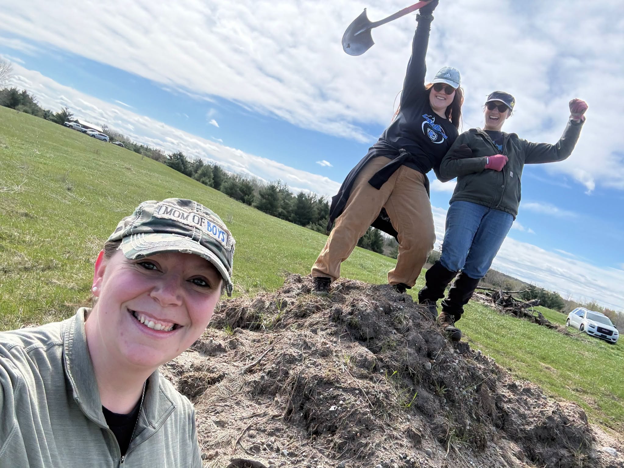 Three people outdoors on a grassy field, one taking a selfie, two standing on a mound, one holding a golf club, sky with clouds.