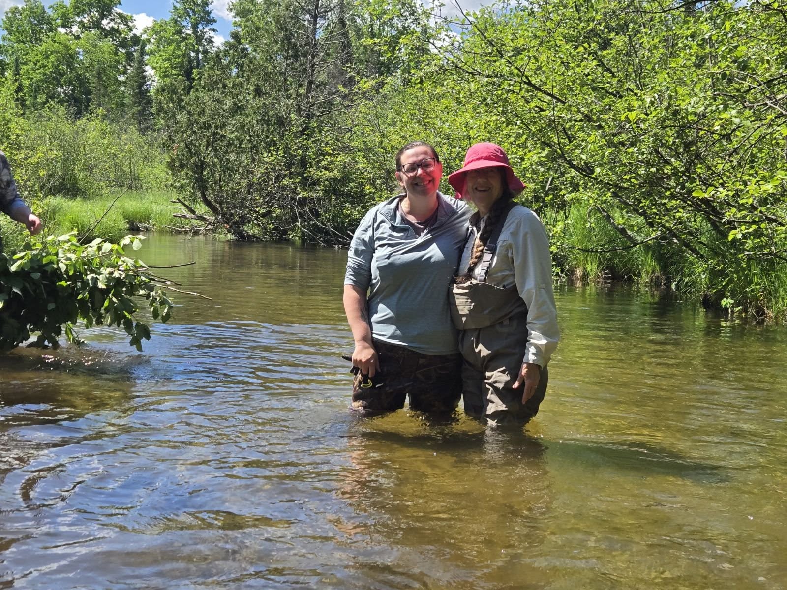 Two people standing in a river with green trees in the background, smiling and wearing outdoor clothing and hats.