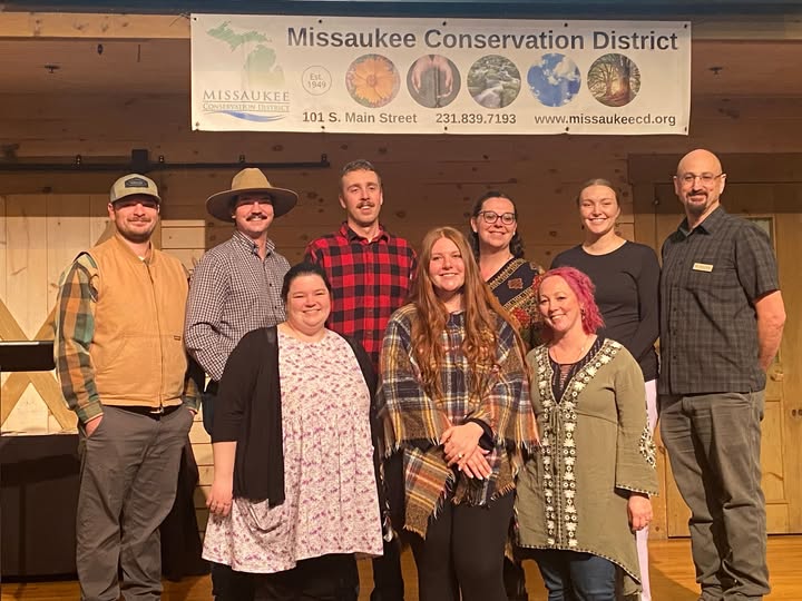 Group of nine people standing in front of a Missaukee Conservation District banner, smiling outdoors.