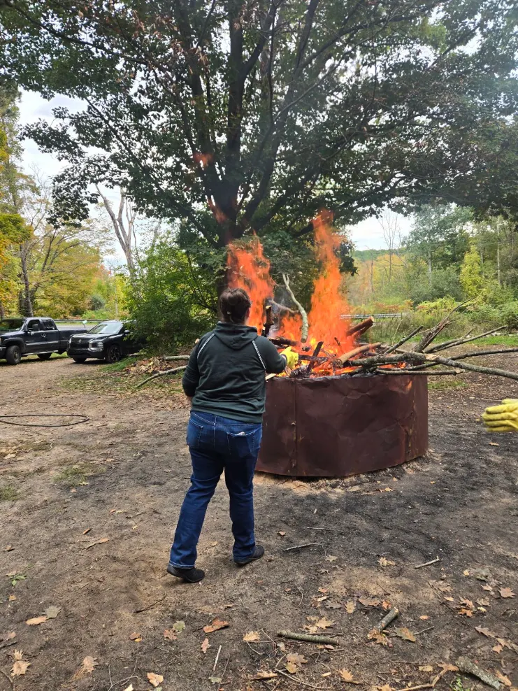 Person standing outdoors near a large fire in a metal container, with trees and parked cars in the background.