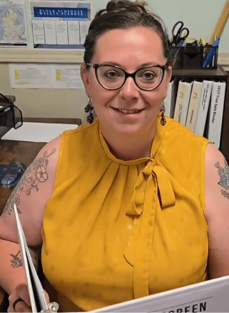 Woman with glasses and earrings, wearing a yellow sleeveless top, sitting at a desk in an office.
