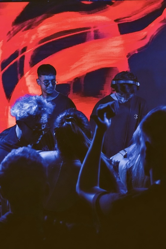 Group of people in a dark setting with red and blue lighting, some wearing glasses, in a club or concert environment.