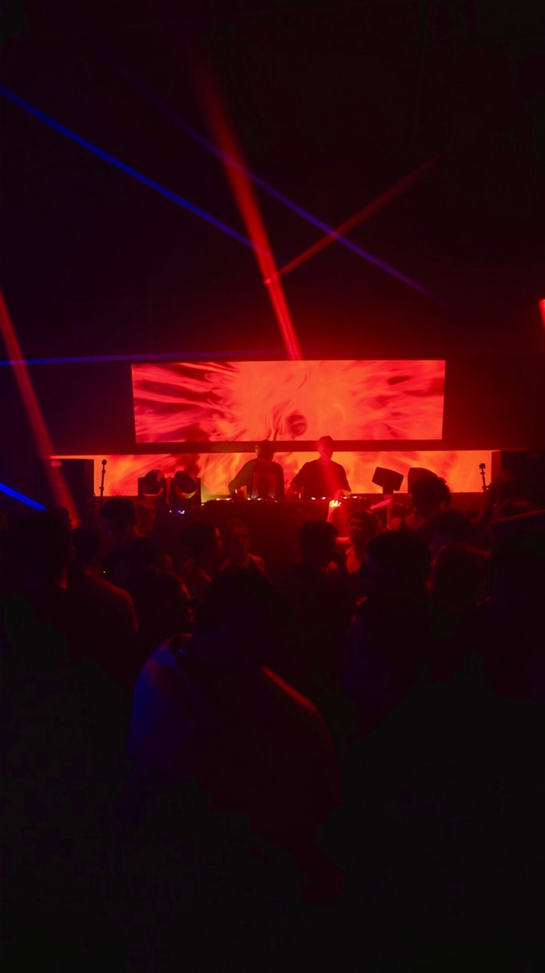 Concert stage with red and blue laser lights and a large screen, audience in foreground, dark environment.