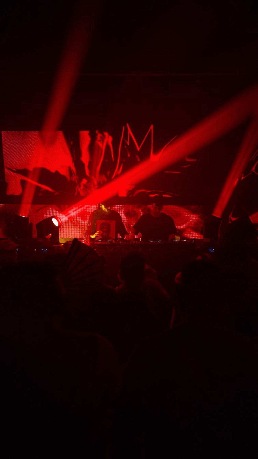 Concert stage with red lighting, screens, and silhouettes of audience members, dark atmosphere, energetic scene.