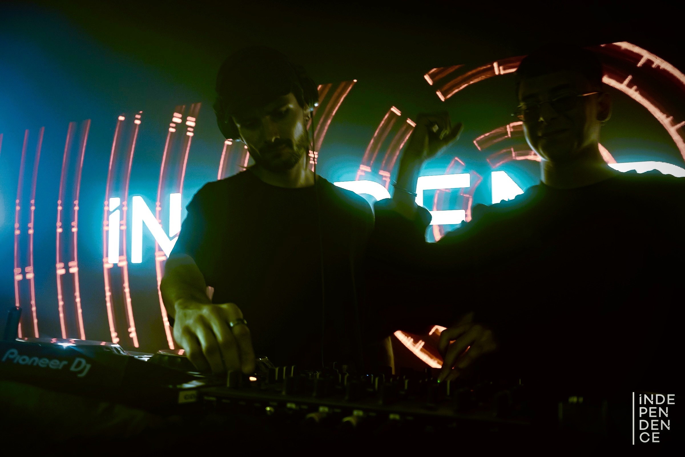 Two DJs perform in front of colorful neon lights, with one adjusting equipment and the other looking on, in a dark setting.