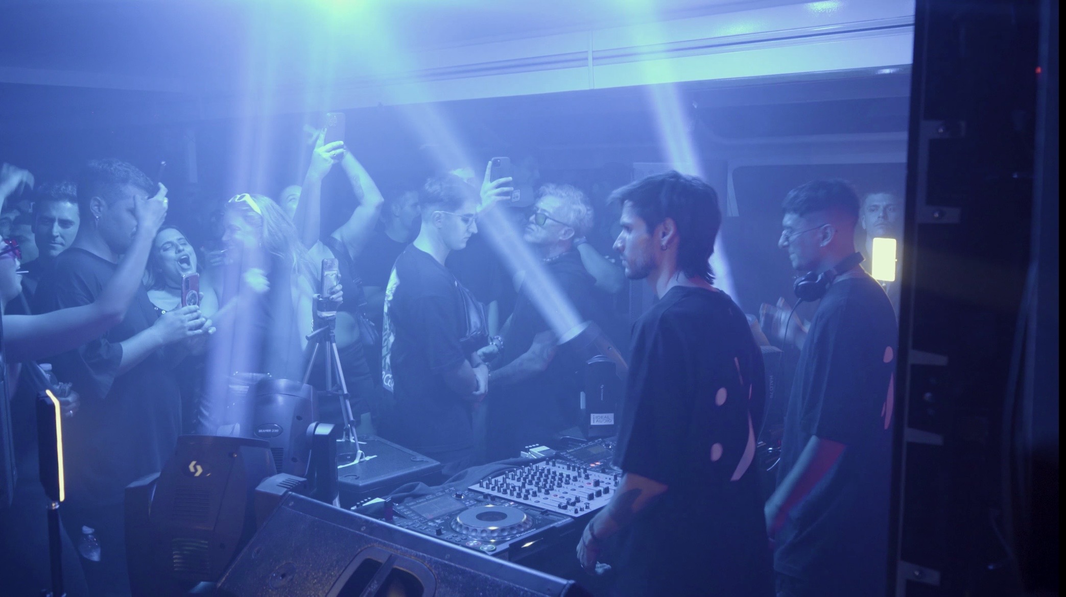 People dancing and DJ playing music in a club with blue lighting, some with hands raised, lively atmosphere.