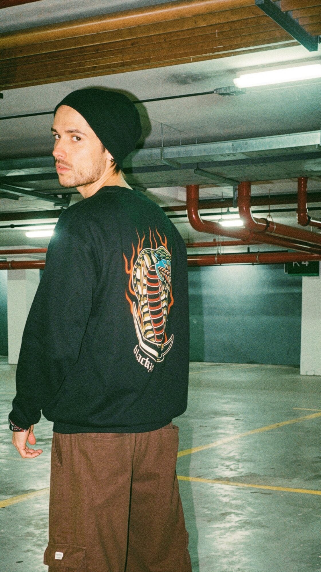 Young man wearing a black beanie and jacket with a colorful design, standing in an underground parking garage.