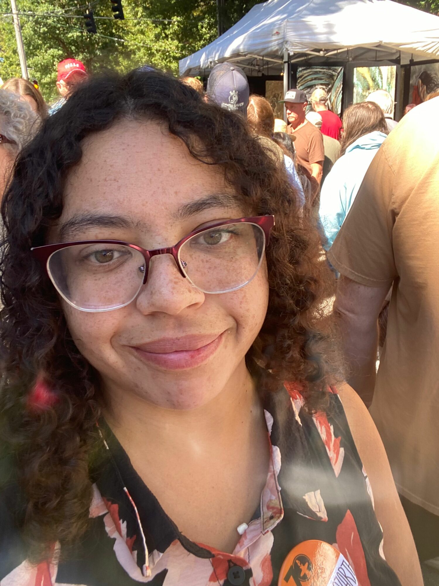 Woman with curly hair and glasses smiling outdoors, people in background, sunny day.