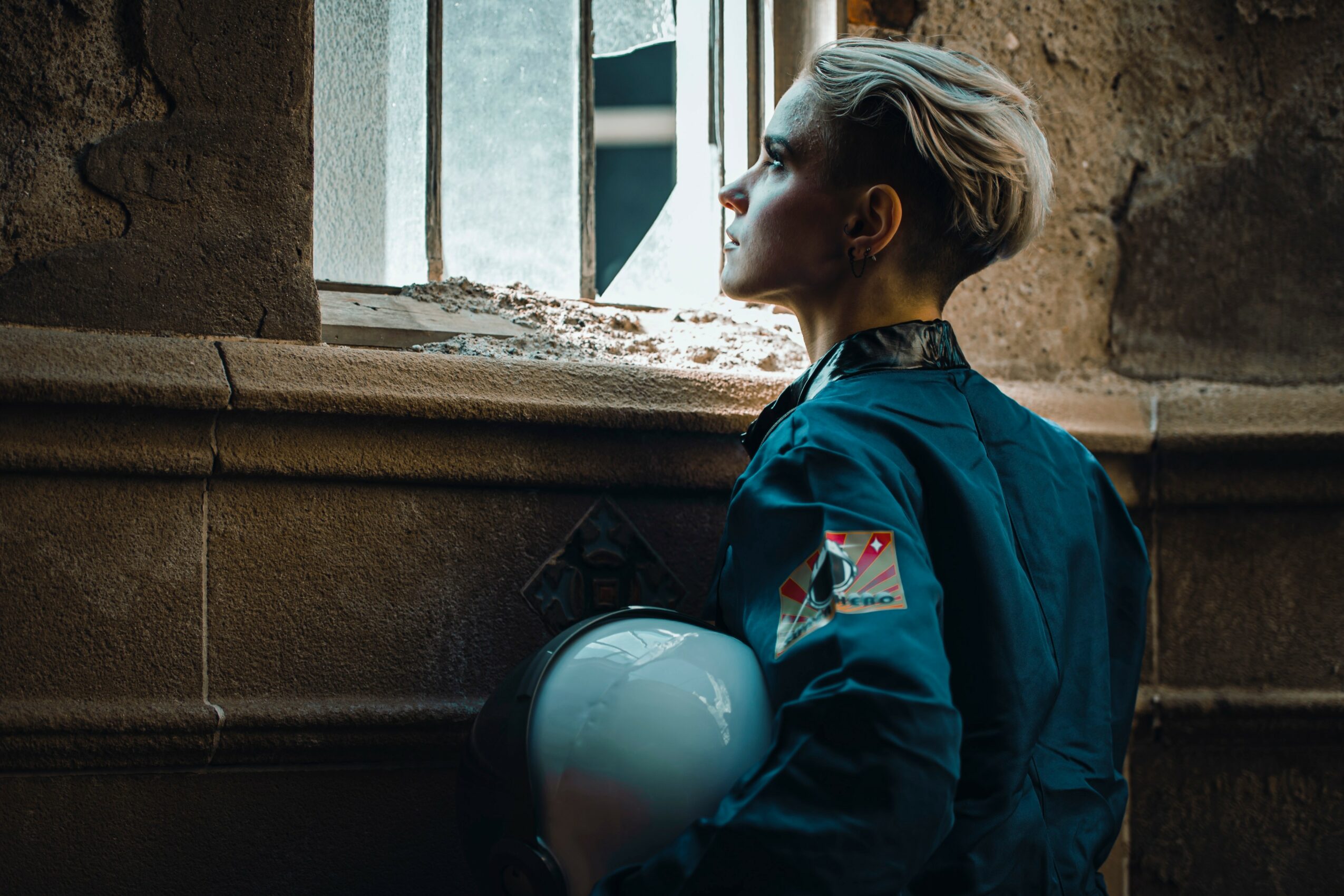 Woman in uniform holding helmet, looking out of a window in a stone building, with light coming through the window.
