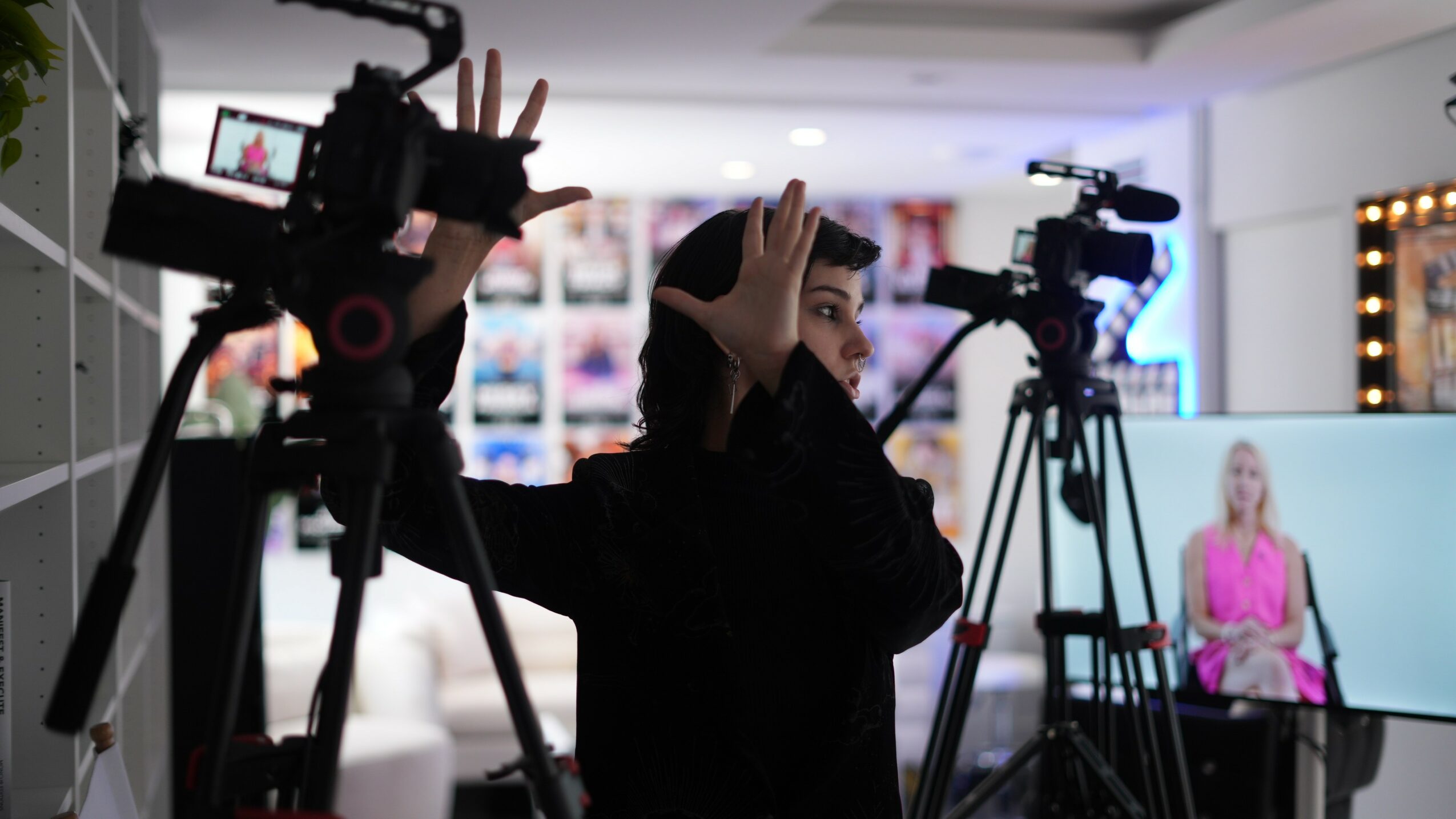 Person operating camera equipment in a studio with a woman in pink on a screen in the background.