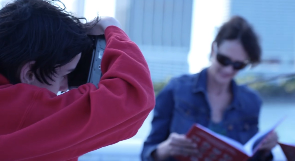 Person taking a photo of a woman reading a book outdoors, both wearing sunglasses, with buildings in the background.