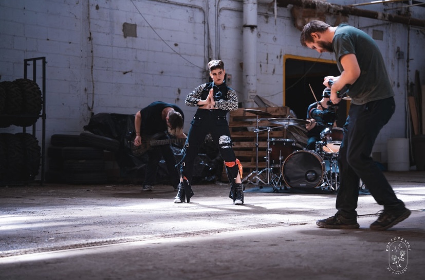 Two children and an adult in a warehouse-like space with drums and tires, engaging in an activity. Dim lighting and industrial background.