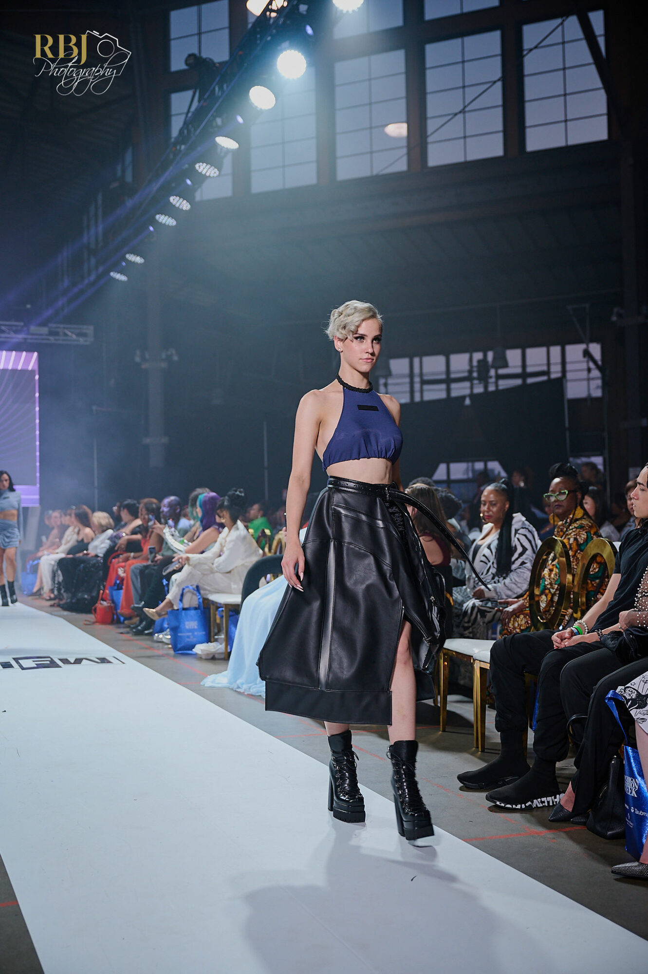 Model walking on runway in a dark blue crop top and black skirt with audience seated on sides, industrial-style venue.