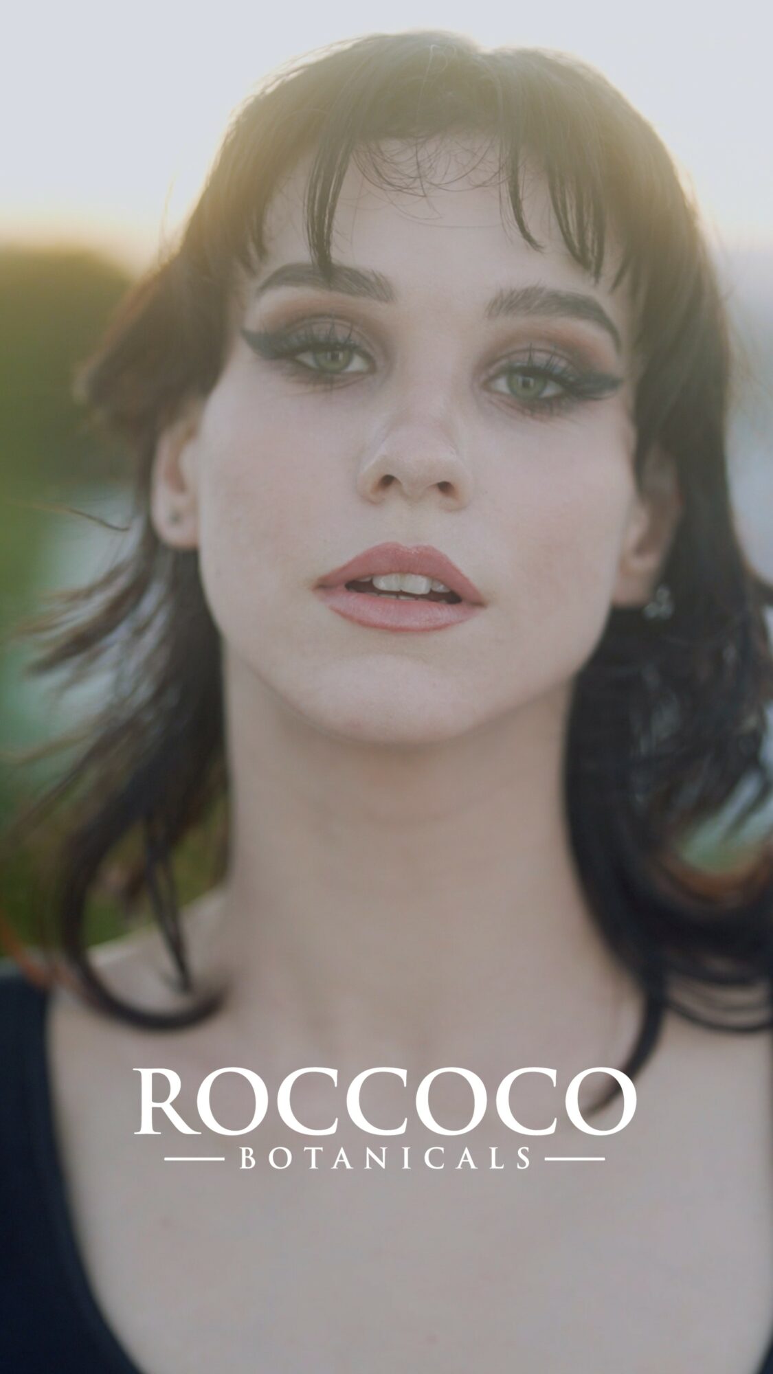 Close-up of a woman with dark hair and makeup, outdoors with blurred background, and text 'ROCCOCO BOTANICALS'.