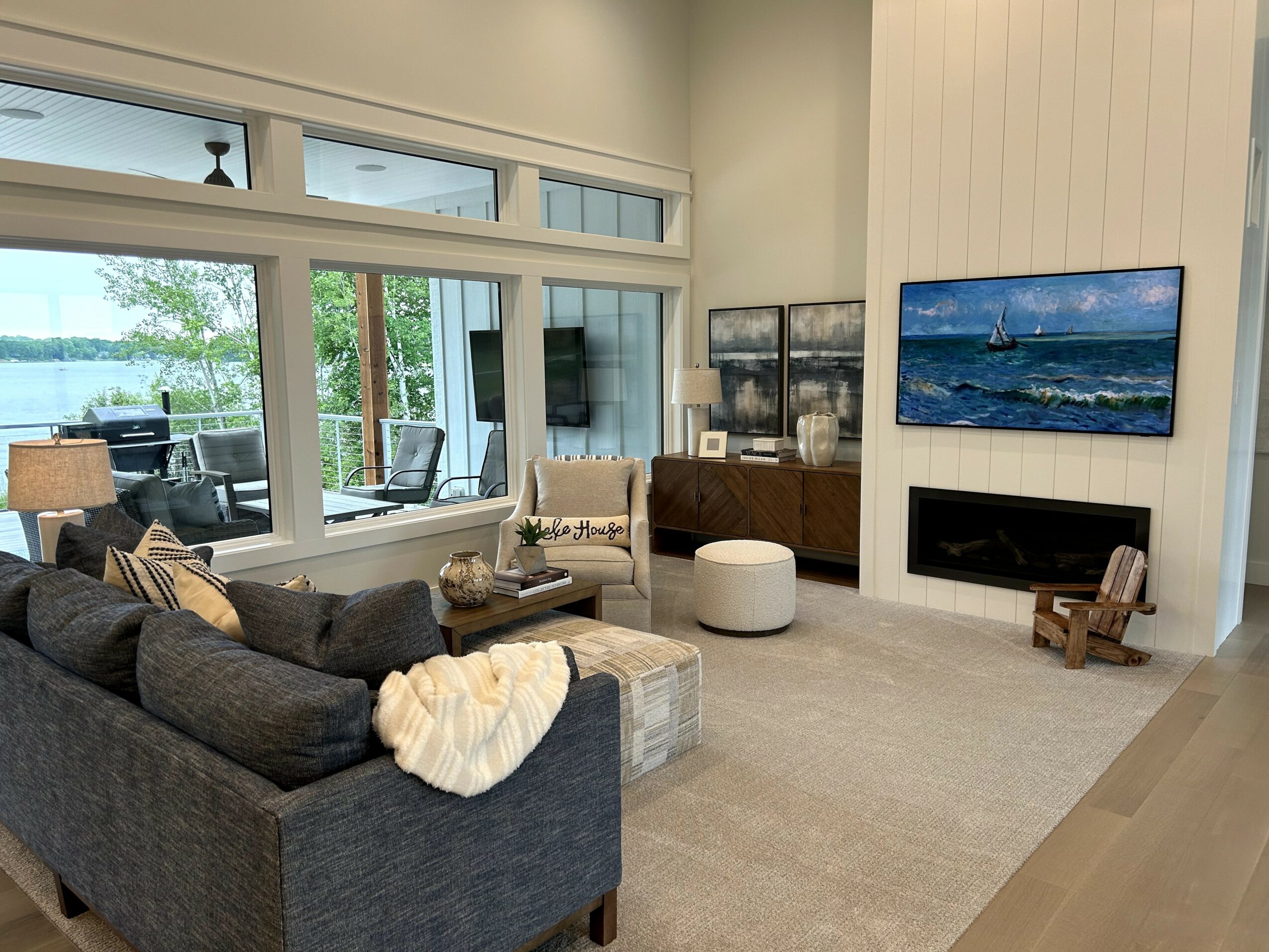 Living room with large windows overlooking water, sofa, armchair, fireplace, and wall-mounted TV, bright and modern interior.