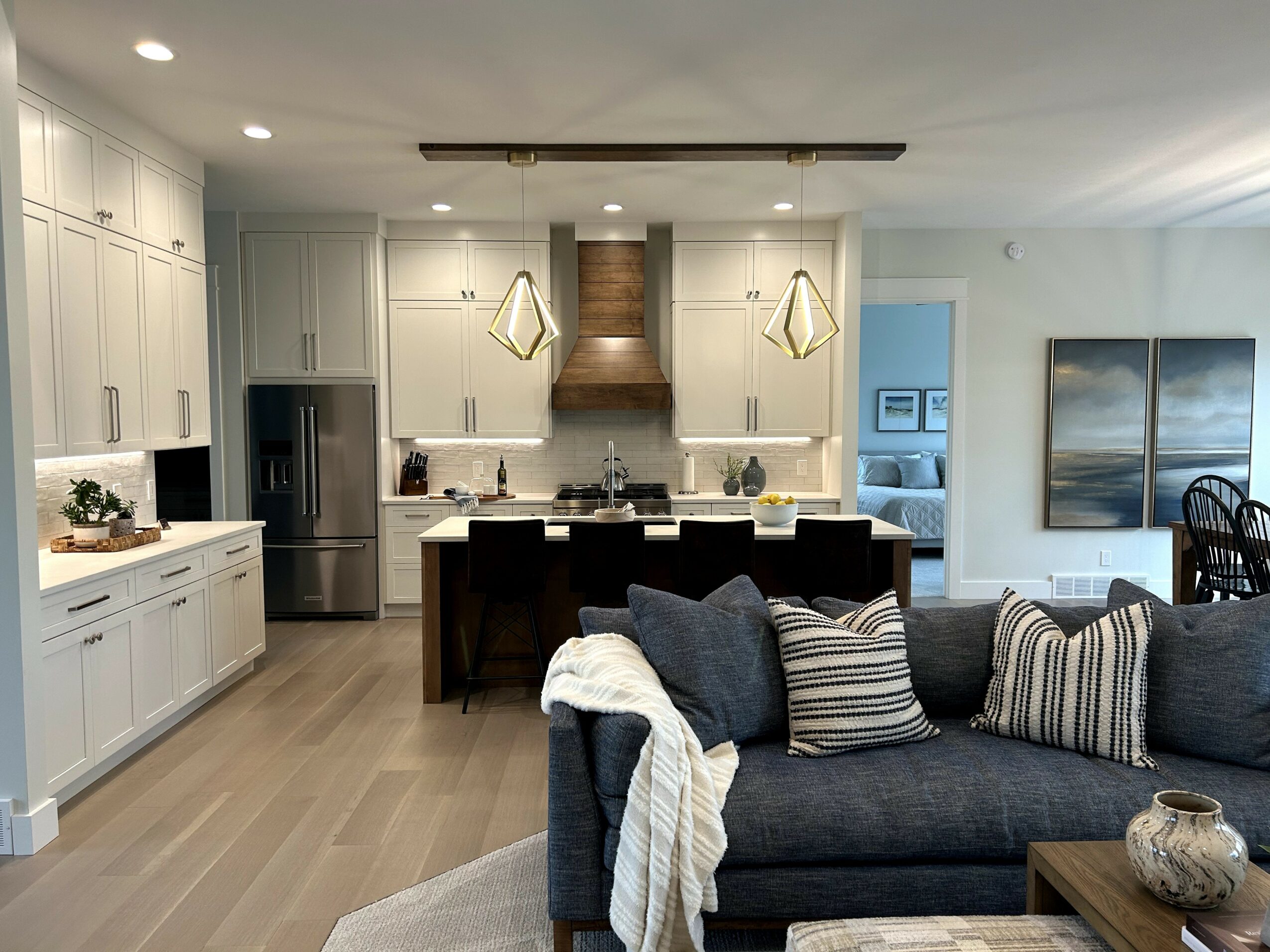 Living room with dark sofa, striped pillows, wooden coffee table, and kitchen in background with white cabinets and stainless steel appliances.