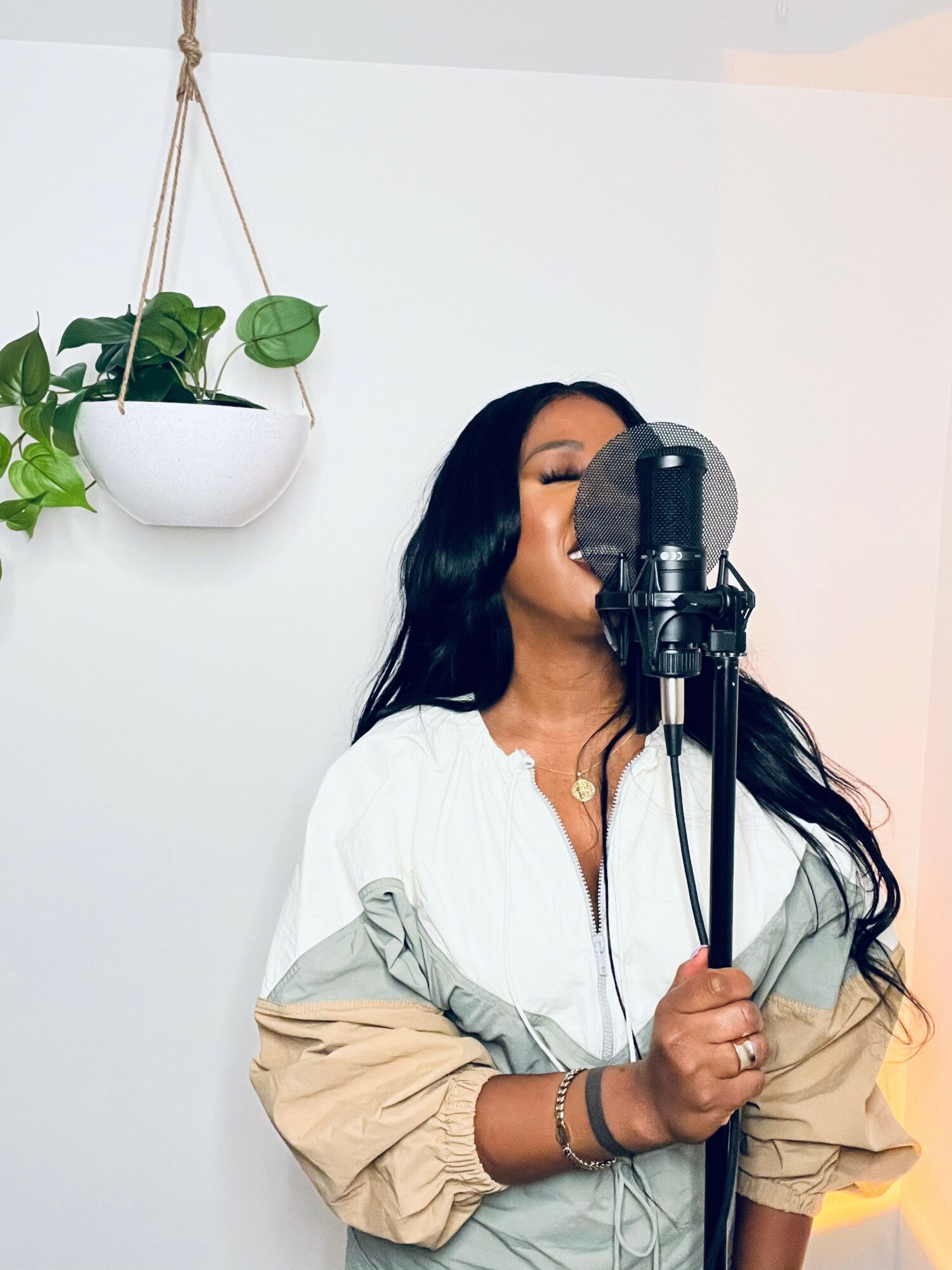 Woman with long dark hair singing into a microphone, wearing a white and beige jacket, in a room with a hanging plant.