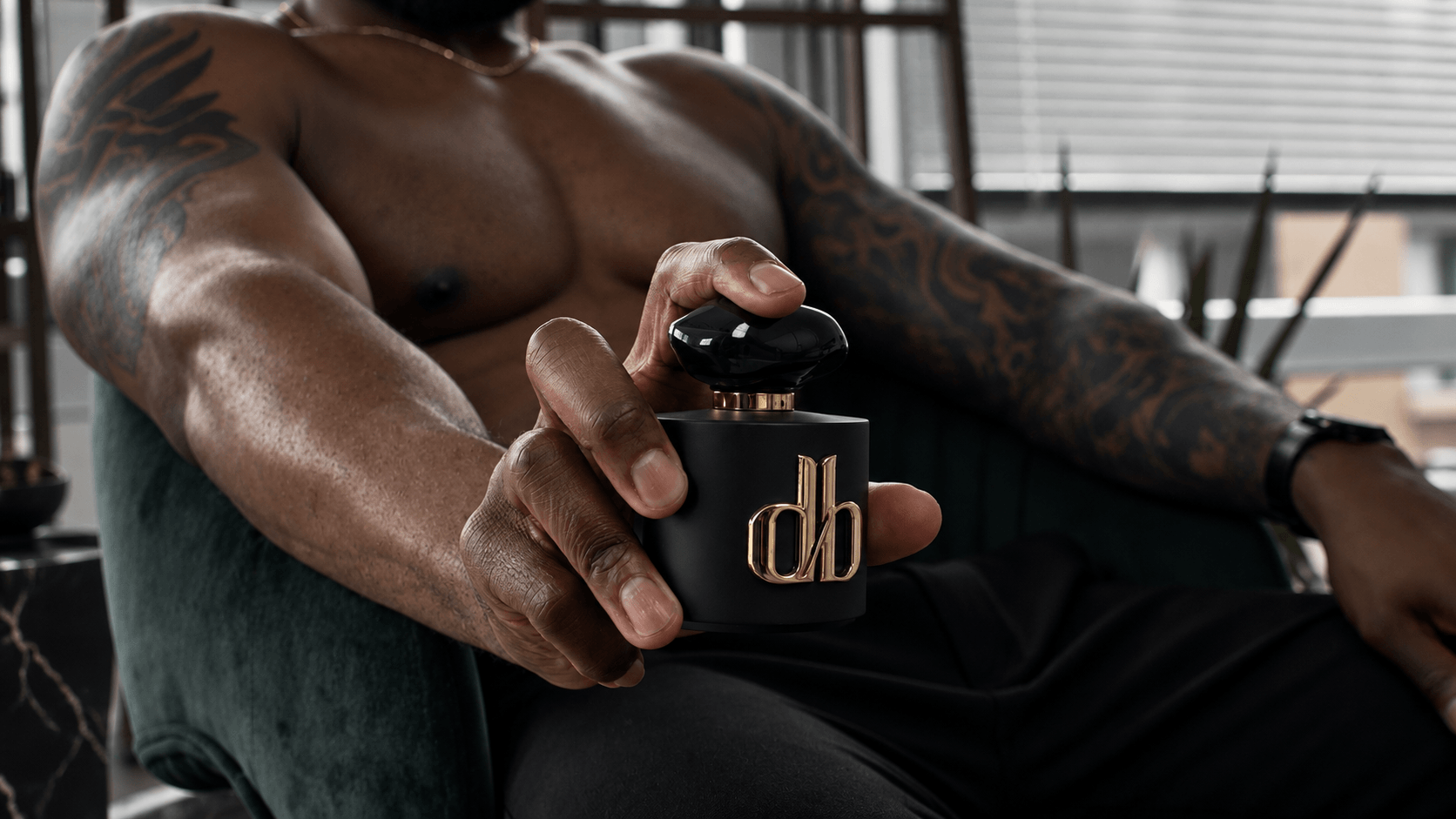 A shirtless person with muscular arms holds a black perfume bottle with gold lettering, sitting indoors near a window.
