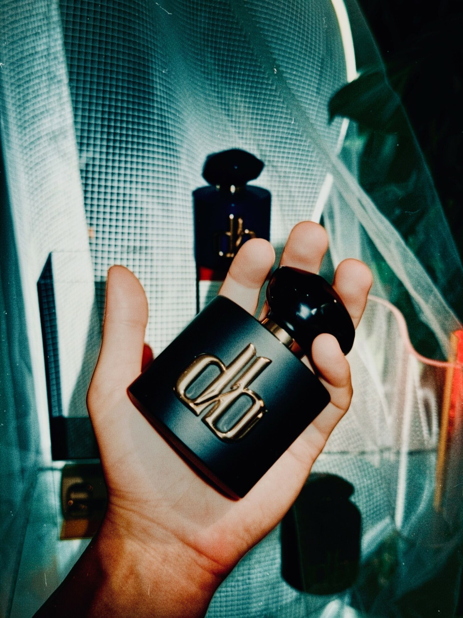 Hand holding a black perfume bottle with gold accents, against a reflective surface and textured background.