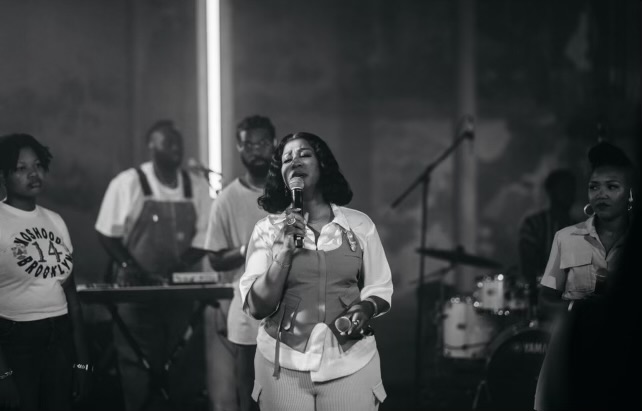 A woman singing into a microphone on stage with musicians in the background, in black and white.