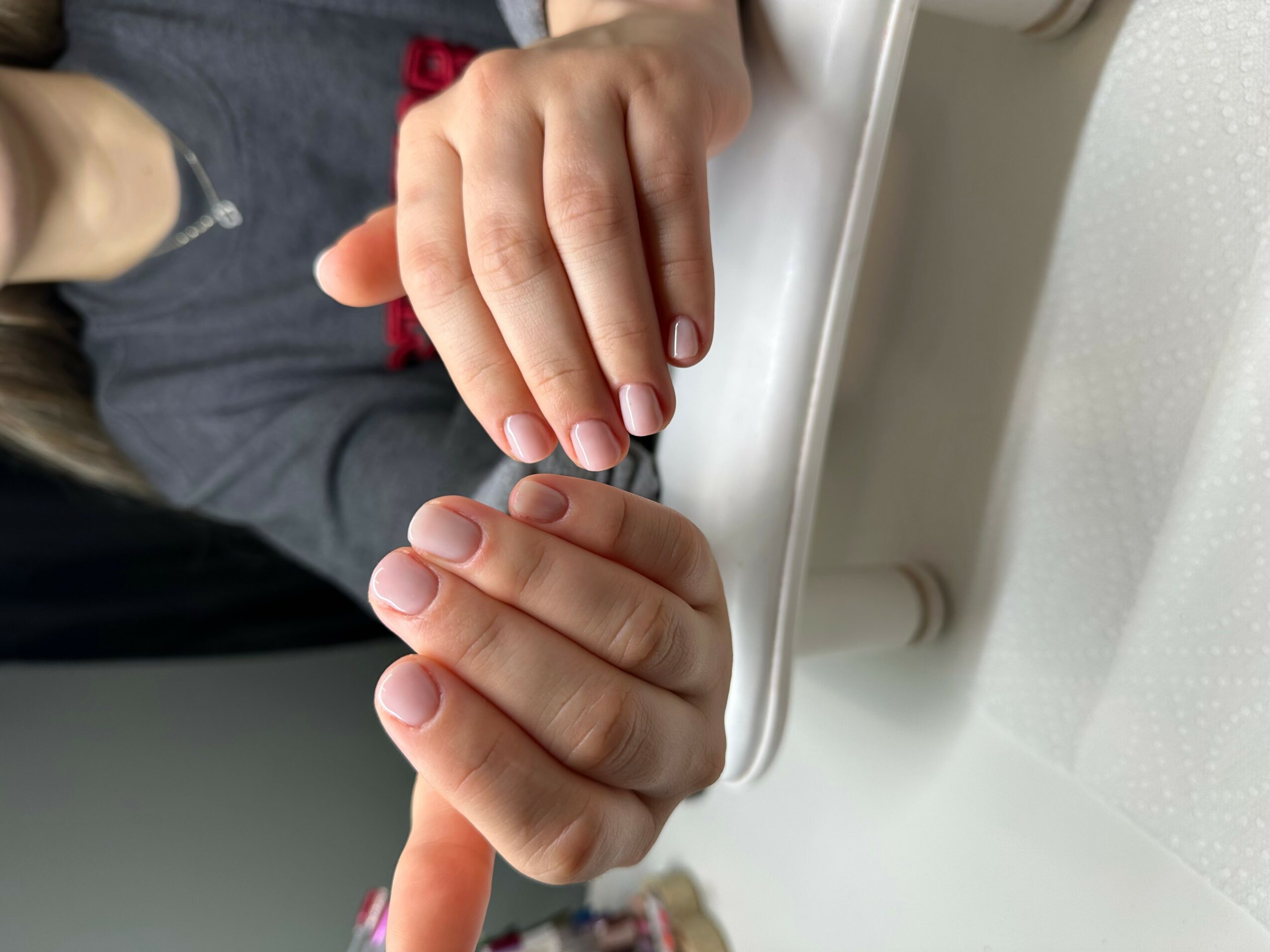 Two hands, one with fingers touching the other, resting on a white surface, with a person wearing a gray shirt in the background.