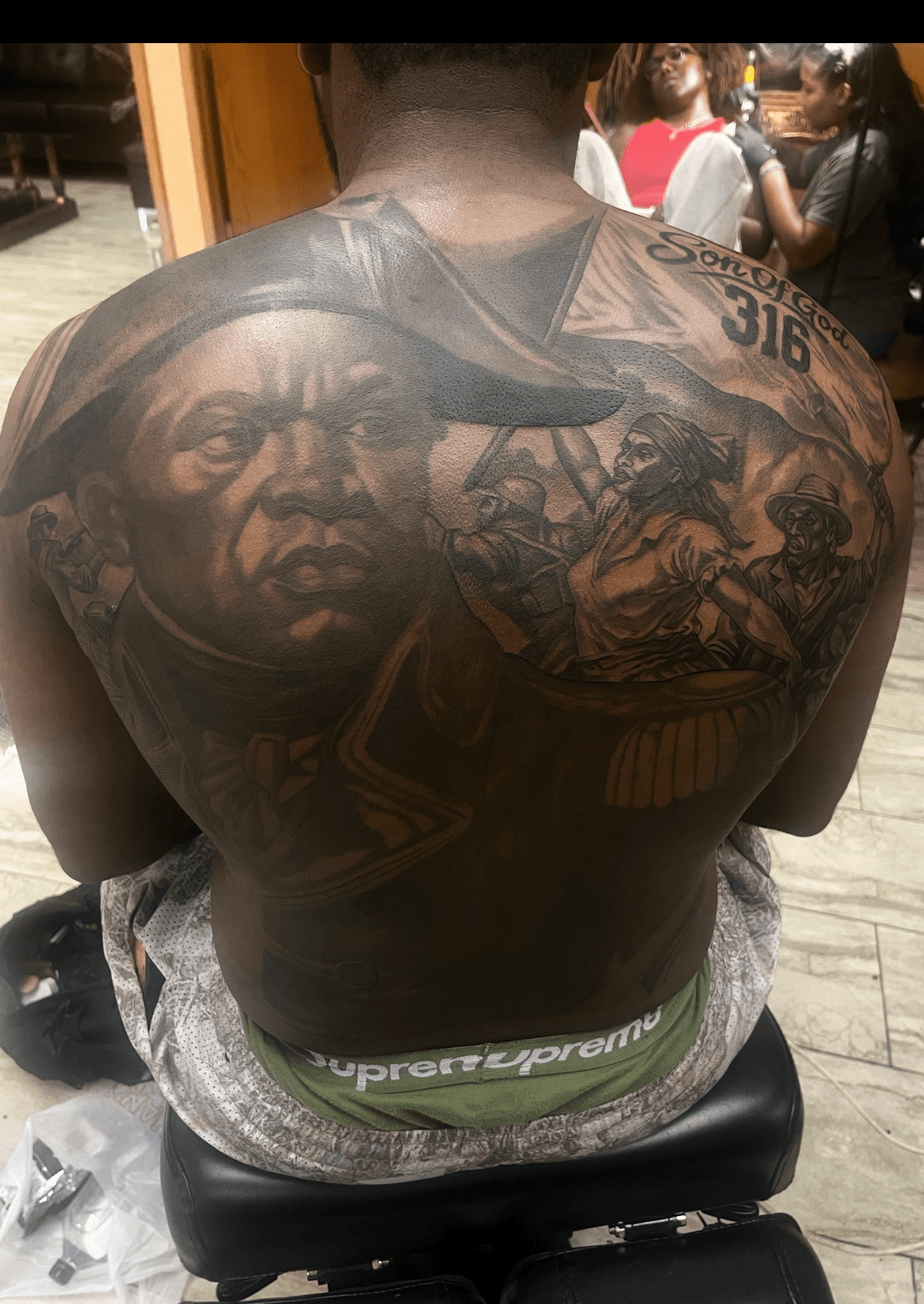 Back of a person with detailed black and gray tattoos, including a portrait, text, and various images, seated indoors.