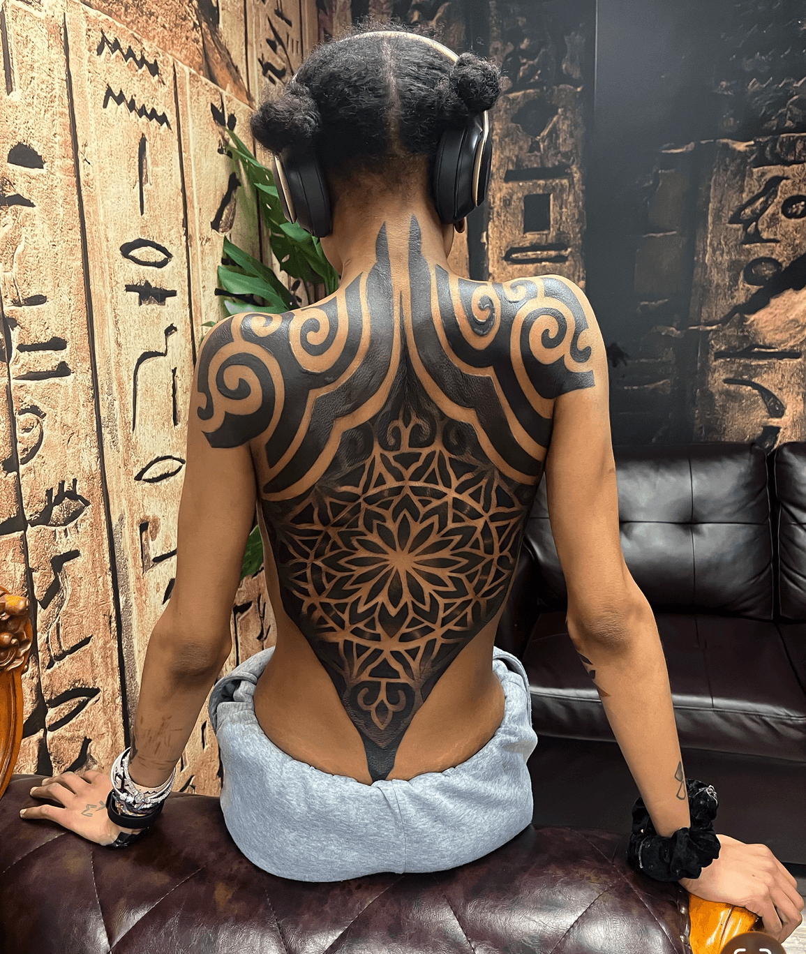 Back view of a person with intricate black tattoo covering shoulders and back, wearing headphones and gray pants, sitting on a leather surface.