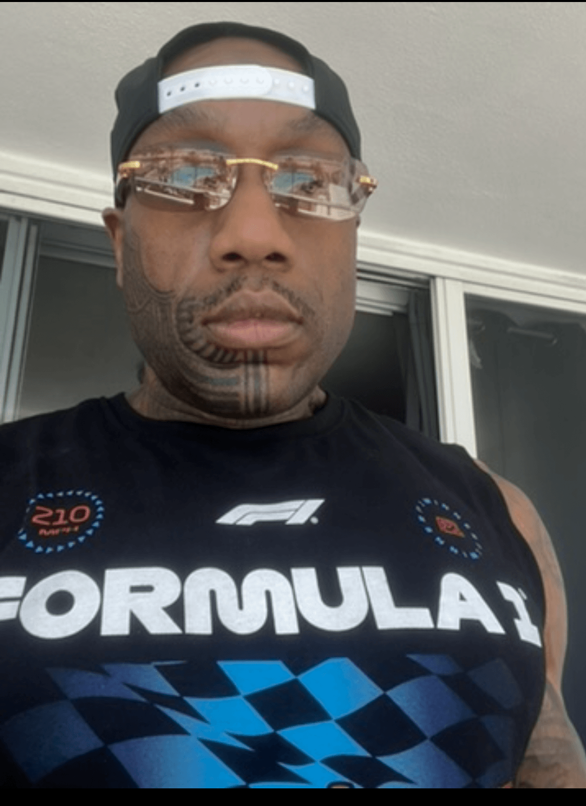 Man wearing glasses, a black cap backwards, and a Formula 1 shirt, standing outdoors near a window.