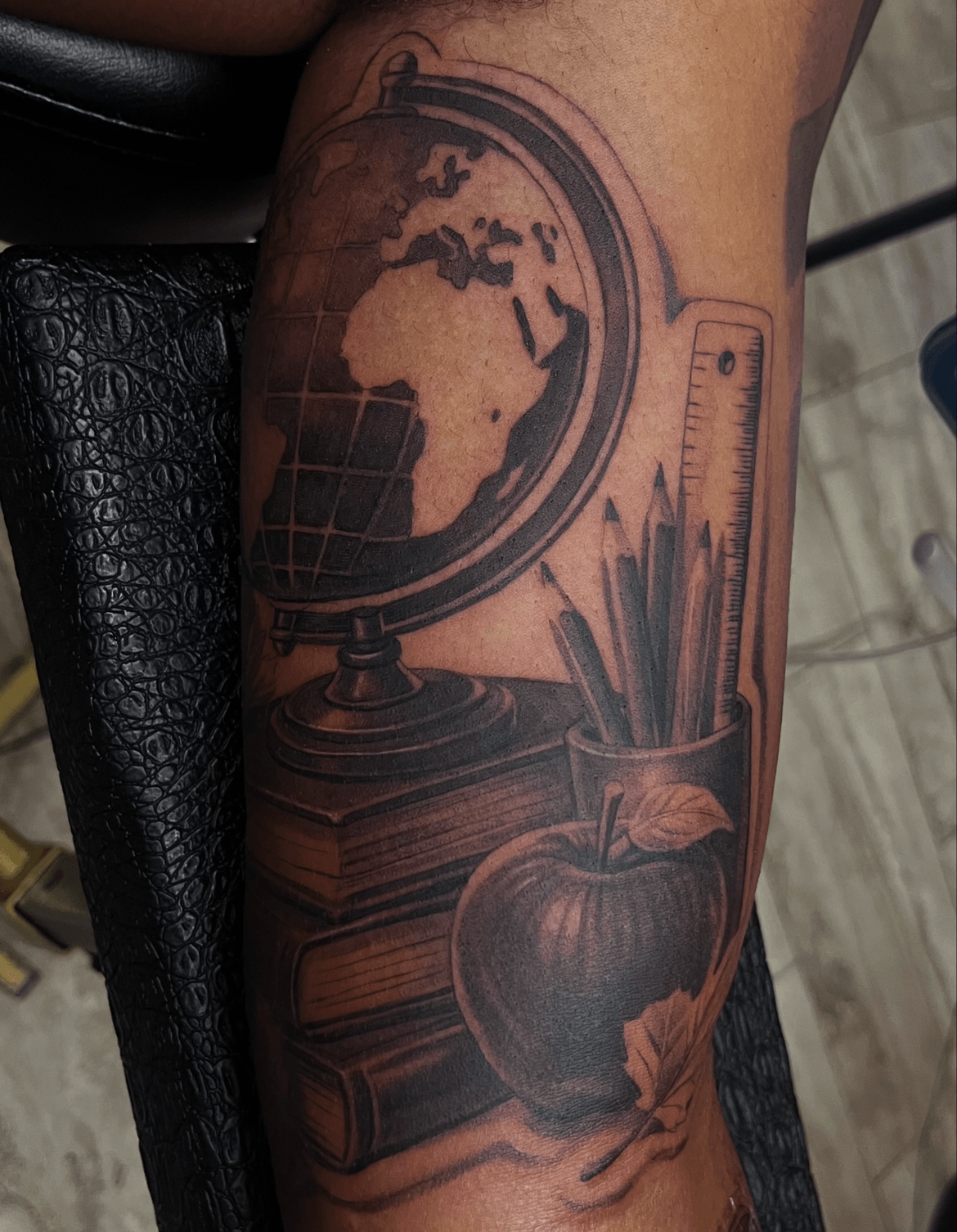Tattoo of a globe, books, and a container with pencils on a person's arm, with detailed shading and line work.