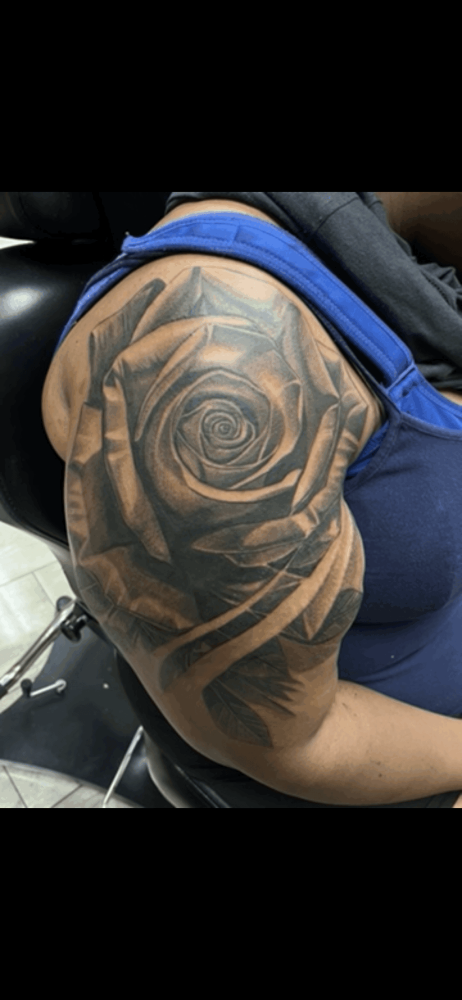 Close-up of a person's shoulder with a detailed black and gray rose tattoo, wearing a blue shirt.