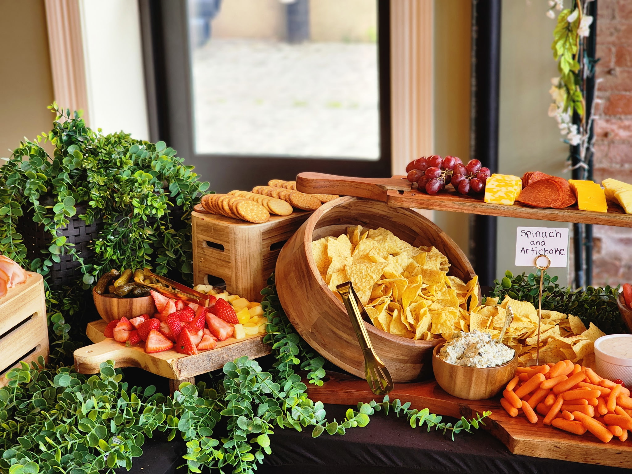 Assorted snacks and vegetables on a table near a window, including chips, sliced strawberries, carrots, and cheese, with greenery nearby.
