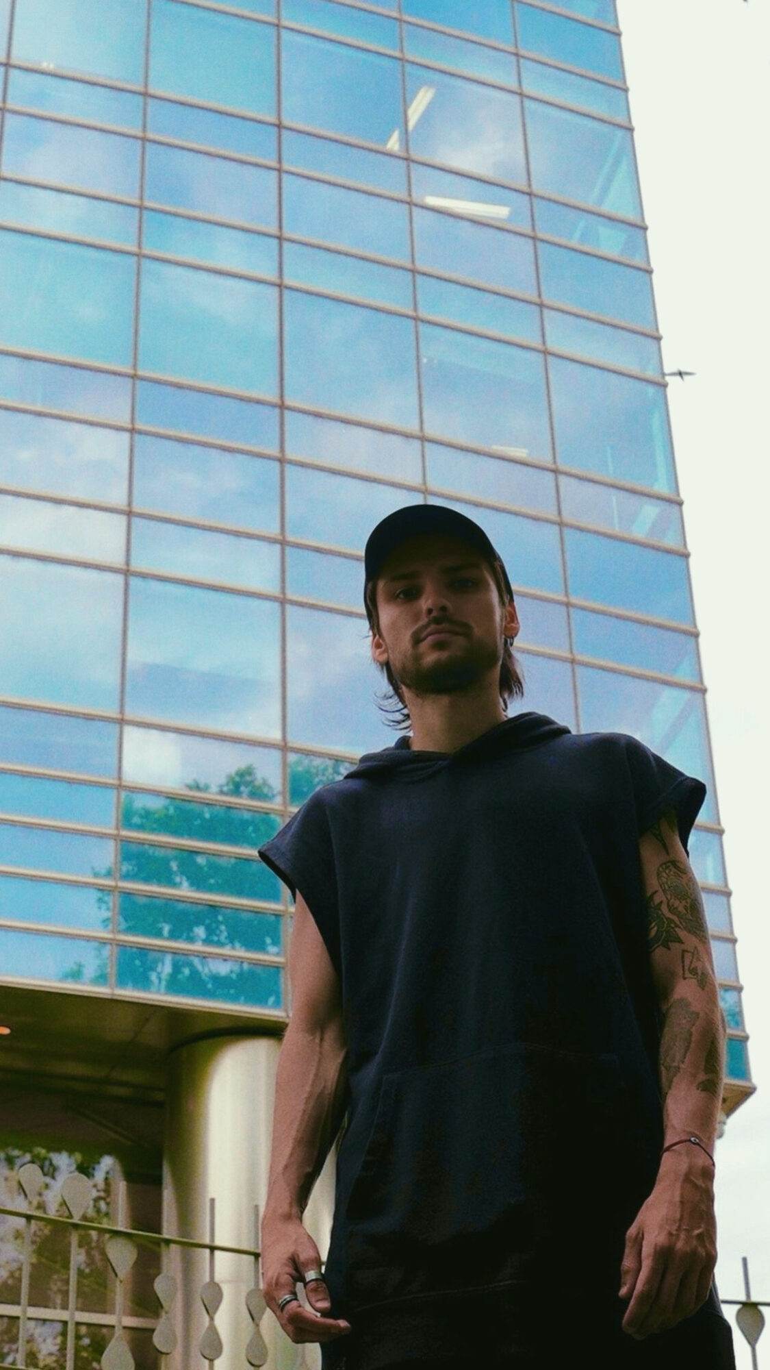 Young man with tattoos wearing a black shirt and cap standing outside near a glass building reflecting the sky.