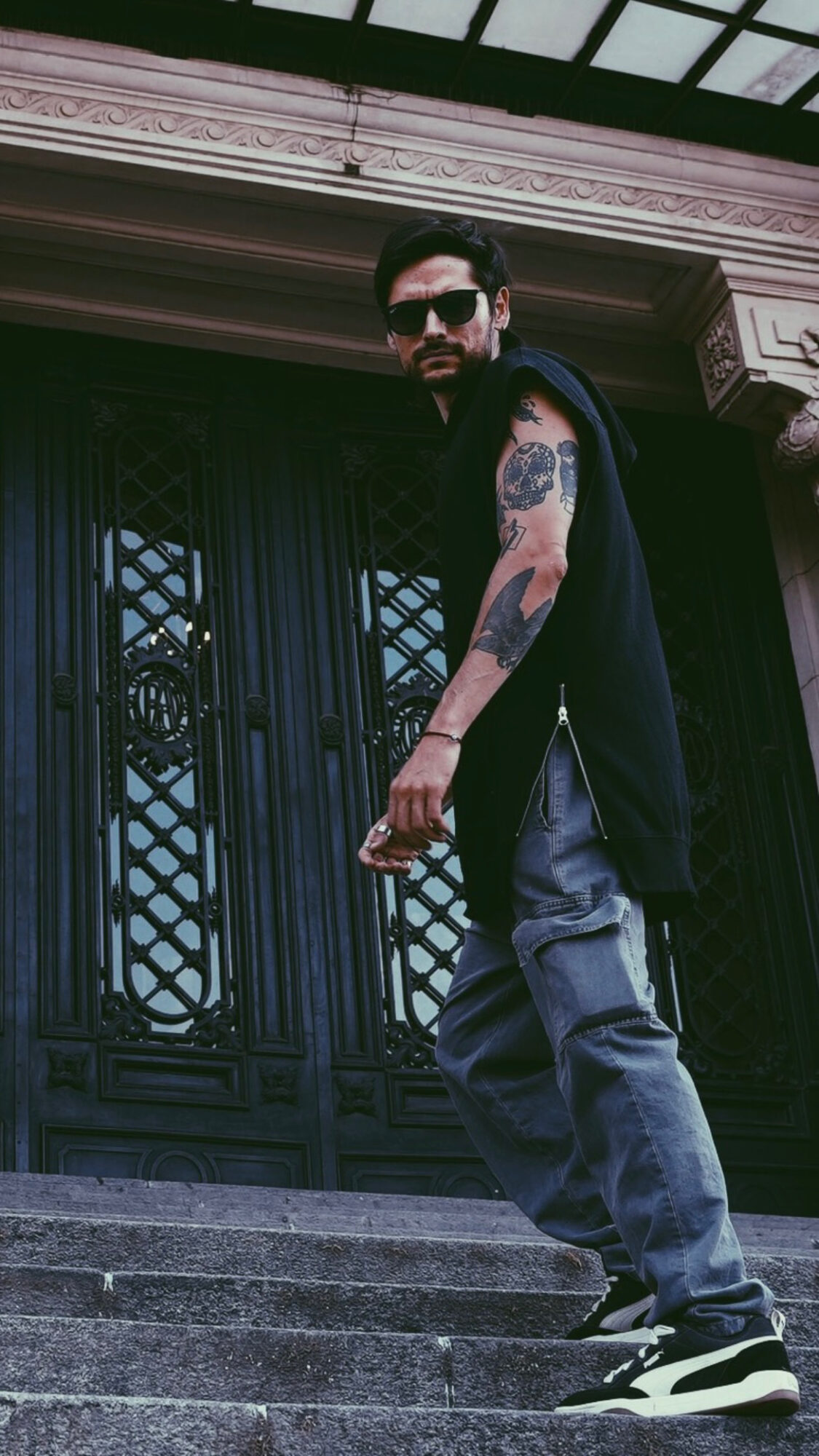 Man with sunglasses, tattoos, black sleeveless shirt, jeans, and sneakers standing on stairs in front of a decorative gate.