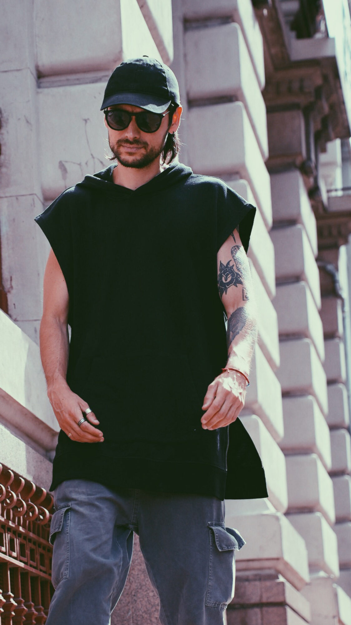 Man with sunglasses, black cap, black sleeveless shirt, tattoos, and gray pants standing outdoors near a building.