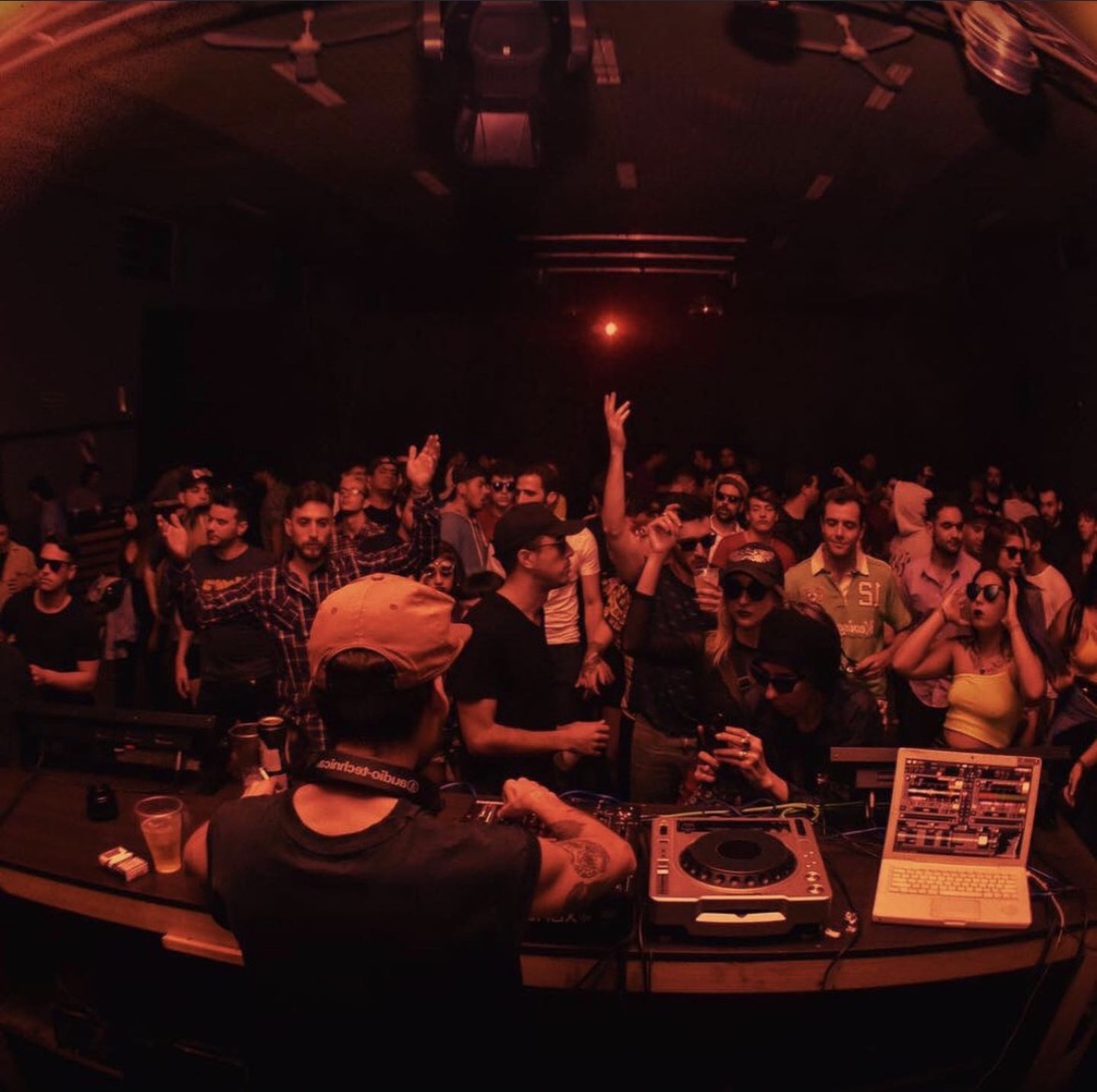 Crowd of people at a DJ event with a DJ in foreground, red lighting, and a laptop, in a dark venue.