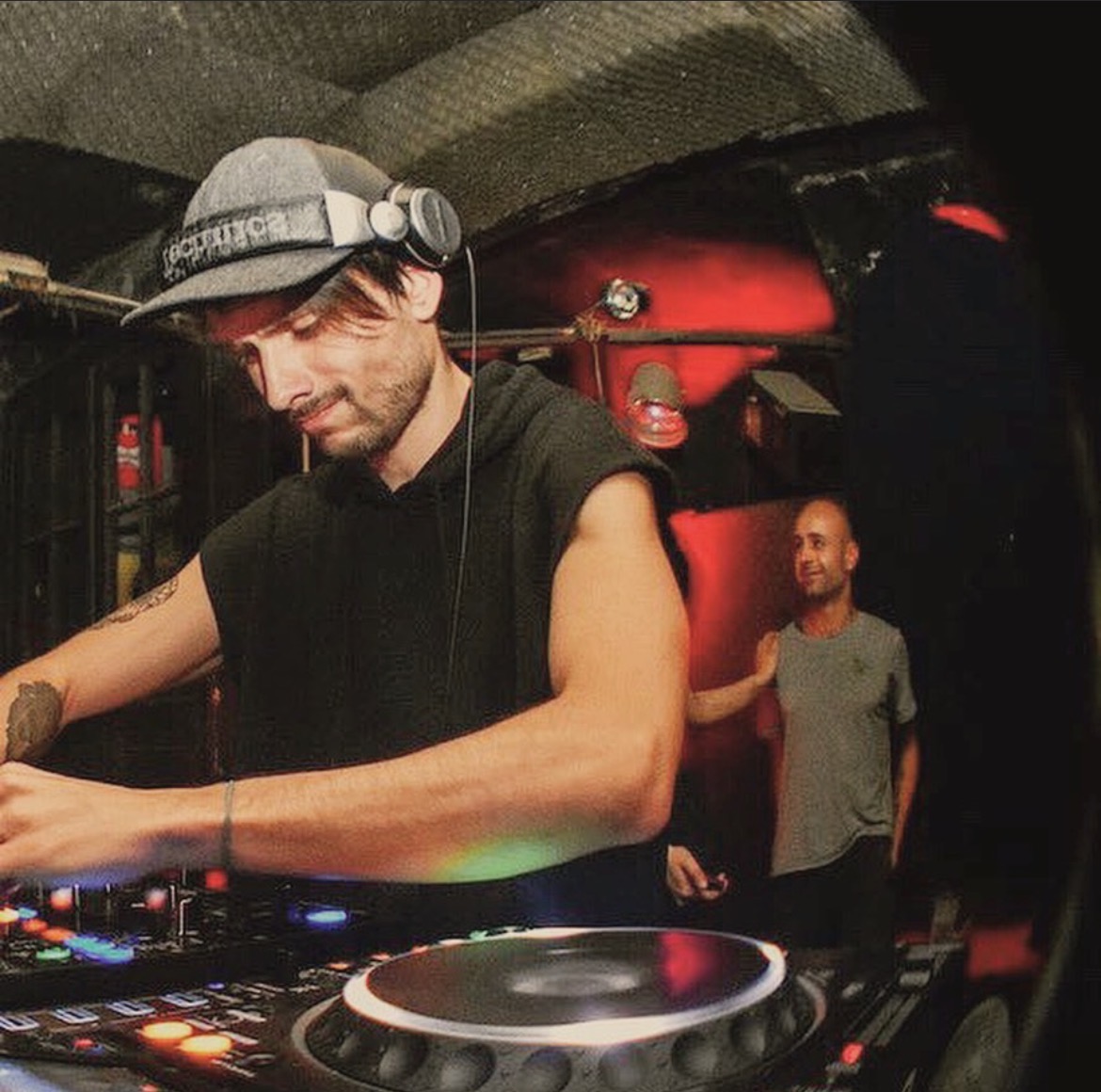 DJ wearing headphones and a cap, operating equipment in a dimly lit club with a person in the background.