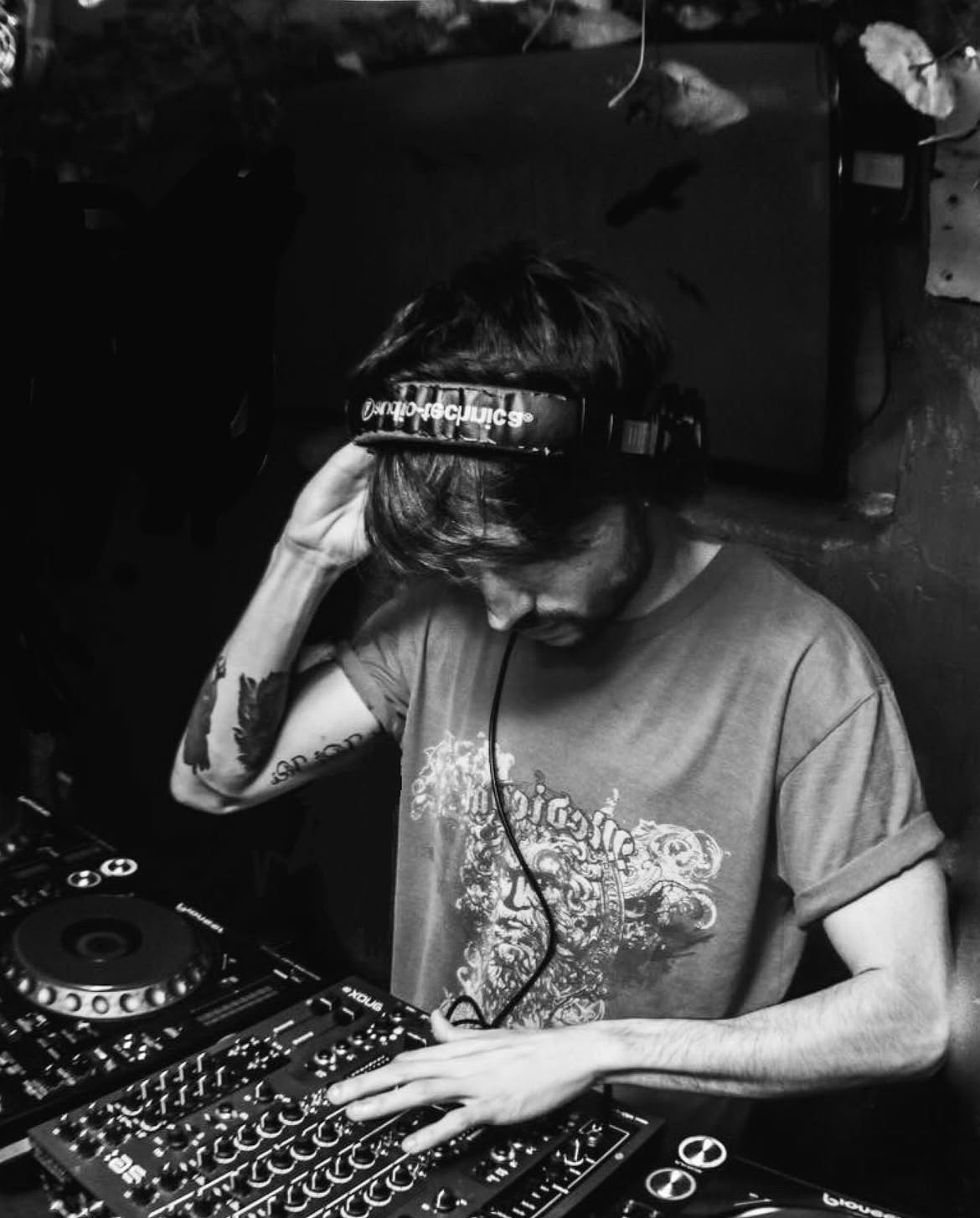 Person wearing headphones, operating DJ equipment, with tattoos on arm, in a dark environment.
